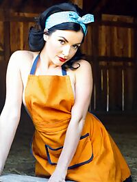 A tousled haired apron clad woman in 1950s style stands in a dusty barn at high noon